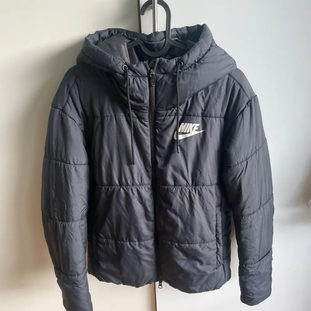 Snygg svart pufferjacka från Nike med huva och dragkedja. Perfekt för kyliga dagar. Jackan har en stilren design med Nike-logga på bröstet.. Takit.