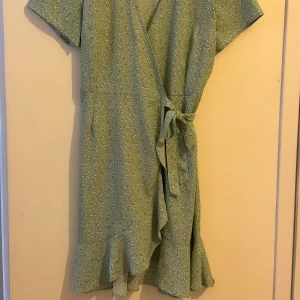 Grön blommig omlottklänning från Bubbleroom storlek M - Söt grön omlottklänning från Bubbleroom med blommigt mönster. Klänningen har korta ärmar och volangdetaljer vid fållen. Perfekt för sommaren!