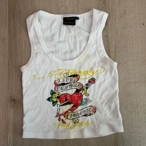 Vit linne från Ed Hardy x Urban Outfitters - Säljer ett coolt vitt linne från Ed Hardy x Urban Outfitters med ett färgglatt tryck av ett hjärta och texten 'Life Before Honor'. Perfekt för en avslappnad stil med en edgy touch.