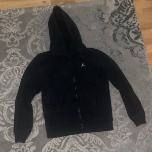 Jordan zip hoodie - Säljer en svart hoodie från Jordan med dragkedja och huva. Den har en liten logga på bröstet och är perfekt för en sportig look. Passar bra till både träning och vardag.