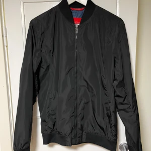 Svart bomberjacka från Zara Man - Snygg svart bomberjacka från Zara Man med dragkedja framtill. Jackan har en klassisk design med ribbad krage, ärmslut och nederkant. Perfekt för en stilren look.