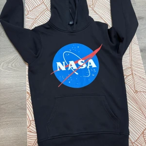 Svart NASA hoodie - Snygg svart hoodie med det ikoniska NASA-logotypen i blått och rött på framsidan. Tillverkad i en mjuk blandning av bomull och polyester. Hoodien är använd en gång.