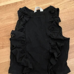 Svart ärmlös topp från H&M - Snygg svart ärmlös topp från H&M med volangdetaljer och broderade mönster längs sidorna. Kommer ta av allt hundhår sedan orkade bara inte göra det nu🤭