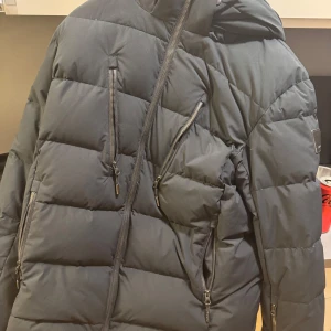 Svart pufferjacka med huva - Snygg svart pufferjacka med huva och dragkedjor på bröstet. Perfekt för kyliga dagar med sin quiltade design som håller värmen. Jackan har en stilren look och passar bra till vardags.  Från revolutionrace!  