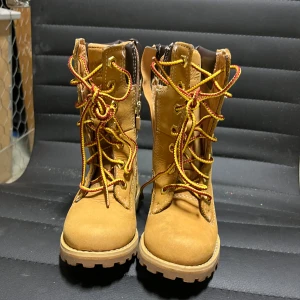 Bruna kängor med snörning och dragkedja - Säljer ett par robusta bruna kängor från Timberland med snörning och dragkedja. De har en slitstark sula och är perfekta för utomhusaktiviteter. Kängorna har en klassisk design med guldfärgade öljetter och flerfärgade snören.