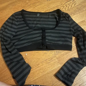 Svart och grå randig bolerotopp - Säljer en svart och grå randig bolerotopp med knappar framtill. Toppen har långa ärmar och en rundad halslinje. Perfekt för att lägga till en extra touch till din outfit.