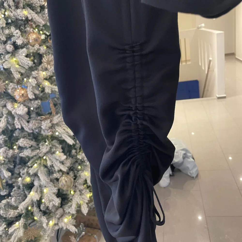 Elegant svart klänning från Twist & Tango med långa ärmar och en stilren design. Klänningen har en dekorativ knytning i sidan för en snygg siluett. Perfekt för en chic look. Bälte medföljer ej.. Mekot.