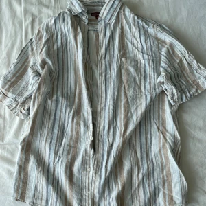 Randig skjorta från Redwood - Säljer en snygg randig skjorta från Redwood i storlek M. Skjortan har korta ärmar och är perfekt för sommaren. Den är i en skön bomullsmix med ränder i beige, vit och grå. Perfekt för en avslappnad stil eller en dag på stan! Frågor? Skriv!