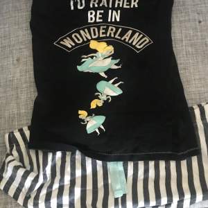 Säljer en svart topp med ett coolt tryck av texten 'I'd Rather Be in Wonderland' och en illustration av en karaktär i blå klänning. Perfekt för dig som älskar unika och lekfulla plagg!