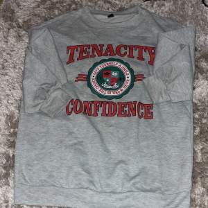 Säljer en grå sweatshirt från SHEIN i storlek XS. Tröjan har tryck med texten 'TENACITY CONFIDENCE' i rött och ett emblem i mitten. Perfekt för en avslappnad stil och passar bra till vardags. Den är i bra skick och superbekväm!