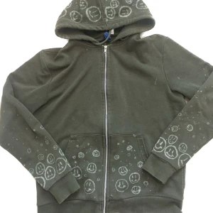 Grön zipup hoodie - En olivgrön zip up hoodie som jag har målat me blekmedel på. Säljer för att den inte kommer till användning. 