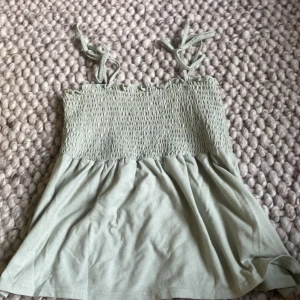 Grön topp från Vero Moda - Säljer en söt grön topp från Vero Moda. Den har smockdetaljer upptill och knytbara axelband. Perfekt för sommardagar och kan enkelt matchas med en kjol eller shorts. Toppen är i ett mjukt och bekvämt material.
