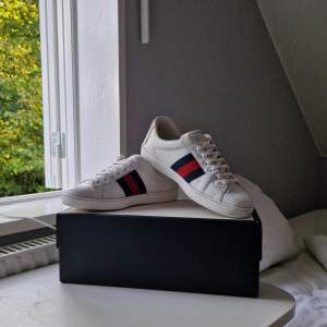 Säljer ett par snygga vita sneakers från Gucci. De har de klassiska ränderna i blått och rött på sidan och en cool tigerdetalj på hälen. Skorna är i skinn och har snörning framtill. Box och kvitto medkommer! Fullt pris ~ 6000KR Mitt pris 2699KR