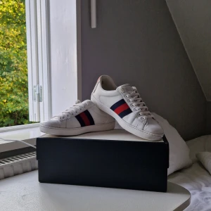 Gucci Ace - Säljer ett par snygga vita sneakers från Gucci. De har de klassiska ränderna i blått och rött på sidan och en cool tigerdetalj på hälen. Skorna är i skinn och har snörning framtill. Box och kvitto medkommer! Fullt pris ~ 6000KR Mitt pris 2699KR