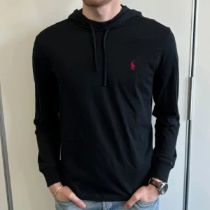 Svart hoodie från Ralph Lauren - Säljer en stilren svart hoodie från Ralph Lauren med det klassiska röda logotypen på bröstet. Tröjan har långa ärmar och en bekväm passform, perfekt för både vardag och avslappnade tillfällen. 
