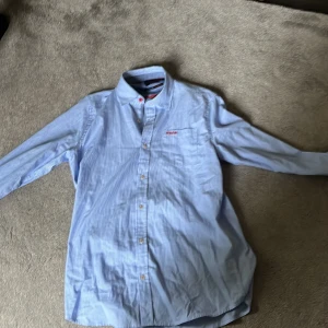 Blå och vit randig skjorta från NZA New Zealand Auckland - Säljer en snygg blå och vit randig skjorta från NZA New Zealand Auckland. Skjortan är i storlek S och har långa ärmar. Den är gjord av bomull och har knappar i en cool röd färg med text. Perfekt för både vardag och lite finare tillfällen!