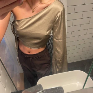 Guldig oneshoulder topp - Säljer en superfin guldig/beige oneshoulder topp med knytning i sidan. Den är croppad och har en lång ärm🤍. Perfekt för festliga tillfällen!