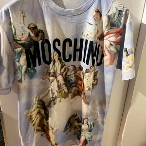 Moschino T-shirt - En väldigt unik Moschino T-shirt med änglar på. Har enbart provat den men aldrig använt tröjan. Skicket är 10/10. Storleken är M/L. Köpt på hemsidan UrbanModern. T-shirten köptes för 4000 kr. 