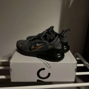 Nike Air Max 270 i grått och svart med orange detaljer  - Säljer ett par Nike Air Max 270 i grått och svart. Skorna har en snygg design med orange detaljer och en stor luftig sula för extra komfort. Perfekta för både vardag och träning. De är i mycket bra skick dock som man ser på bilden ett jätte jätte litet hål, skorna kommer i original kartong.