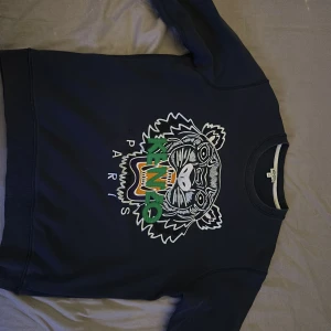 Mörkblå sweatshirt från Kenzo - Säljer en snygg mörkblå Kenzo sweatshirt med ett coolt tigertryck och texten 'Paris' på framsidan. Tröjan har långa ärmar och en ribbad krage. Perfekt för en avslappnad stil eller att bära till vardags. Passar bra till jeans eller chinos!