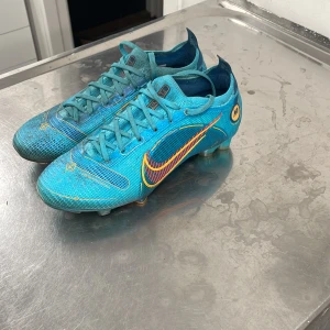 Blå Nike Mercurial fotbollsskor - Säljer ett par snygga blå Nike fotbollsskor i bra skick. De har en cool design med orange detaljer och snörning. Perfekta för fotbollsträningen eller matchen. Skorna har en tight passform och är gjorda i syntetmaterial för bästa prestanda på planen.