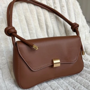 Brun axelväska med gulddetaljer - Säljer en snygg brun axelväska med stilrena gulddetaljer. Väskan har en elegant design med en knäppning framtill och en axelrem med knutdetaljer. Perfekt för både vardag och fest! Oanvänd