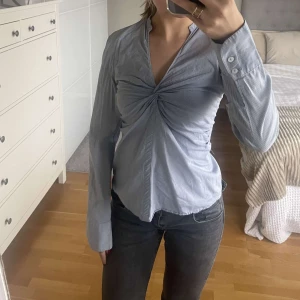 Blå randig blus med knyt - Säljer en snygg blå randig blus med en unik knytning framtill. Blusen har långa ärmar och en v-ringad design som ger en elegant touch. Från zara