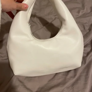 Vit axelväska med dragkedja - Säljer en stilren vit axelväska i syntetmaterial. Väskan har en praktisk dragkedja och en mjuk, rundad form som gör den perfekt för vardagsbruk. Passar till alla säsonger och outfits!     Den har endast används en gång och det är på min bal😍 så nästan en helt ny väska för ett jättebra pris🤩 100kr eller högsta bud men först till kvarn!