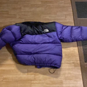 Lila och svart dunjacka från The North Face - Säljer en snygg lila och svart dunjacka från The North Face. Jackan är perfekt för kalla vinterdagar med sin pufferstil och långa ärmar. Den har en dragkedja framtill och justerbara snören i nederkant för bästa passform. Perfekt för både vardag och äventyr! 💜