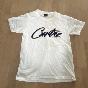 Vit t-shirt med tryck - Säljer en vit t-shirt från Cortez i storlek XS. Den har ett coolt tryck med text och en grafisk design på framsidan. Perfekt för en avslappnad stil eller sommarhäng. T-shirten är i bra skick och gjord av mjukt material.