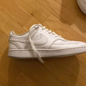 Vita Nike sneakers court vision - Säljer ett par klassiska vita Nike sneakers i skinn. De har en stilren design med snörning och perforerade detaljer på tån för extra ventilation. Perfekta för vardagsbruk och passar till nästan alla outfits. Skorna är i bra skick med lite slitage på sulan.
