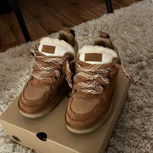 Säljer ett par snygga bruna sneakers från UGG i nytt skick. De har en mysig foder av päls och coola snören i vitt och brunt. Perfekta för höst och vinter med sin varma design. Skorna är i storlek dam 38 och kommer i originalkartong.