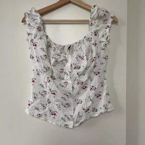Vit blommig blus med volangdetaljer Nelly - Söt vit blus med små röda och gröna blommor från Nelly. Den har volangdetaljer vid axlarna och knappar framtill. Perfekt för vår och sommar! 🌸Inga nopprar eller tecken på föråldrad, utan väldigt fint skick