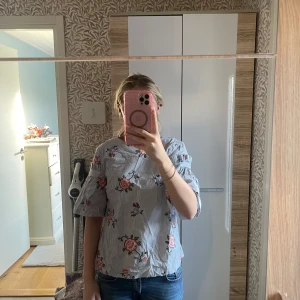 Blommig blus från H&M - Säljer en söt blommig blus från H&M i storlek 34. Den har ett randigt mönster i blått och vitt med vackra blommor i rosa och orange. Blusen har trekvartsärmar och en liten öppning i ryggen. Perfekt för vår och sommar!