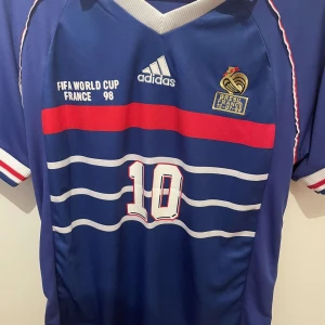 Fotbollströja Frankrike - Säljer en blå fotbollströja från Adidas med korta ärmar. Tröjan har vita och röda detaljer, inklusive ränder och text. Den har nummer 10 och texten 'FIFA World Cup France 98' på framsidan. Baksidan har namnet 'Zidane' och nummer 10. Perfekt för fotbollsfans! Skick 10/10. Aldrig använd. Det är S men stor i storleken så passar M med! Tröjan är äkta. Bara att höra av sig vid minsta fundering!