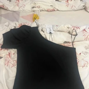 Svart oneshoulder tröja - Snygg svart oneshoulder tröja i nytt skick. Perfekt för festliga tillfällen med sin eleganta design. Tröjan har en volangdetalj vid axeln som ger en extra touch av stil. Passar bra för både sommar och nyårsfester.