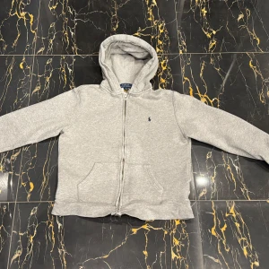 Ralph Lauren kofta  - Säljer en klassisk grå hoodie från Ralph Lauren i storlek L/G (14-16). Den har en dragkedja framtill och en liten broderad logga på bröstet. Perfekt för en avslappnad stil och passar till alla säsonger. Superbekväm och stilren! Nypris:1300kr