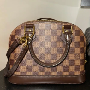 Brun rutig handväska med röd insida - Säljer min LV väska som är som ny. Endast använd ett fåtal gånger, finns certifikat. Säljs för 15 000kr, nypris 19 200kr. Pris går att diskutera vid snabb affär. Mina initialer står på ena sidan som det syns på bild men det går att vänta sida på den så att det inte ska synas