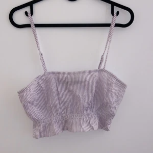 Randig linnetopp - Söt randig linnetopp i ljuslila och vit med smala axelband. Perfekt för sommardagar och kan enkelt matchas med jeans eller shorts. Toppen har en lätt volangdetalj vid nederkanten som ger en extra touch.