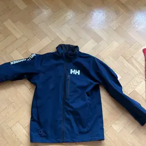 Säljer en snygg blå vindjacka från Helly Hansen. Den har en dragkedja framtill och är långärmad med loggan tryckt på ärmen och bröstet. Perfekt för blåsiga dagar och utomhusaktiviteter. Jackan är i bra skick och redo för nya äventyr!