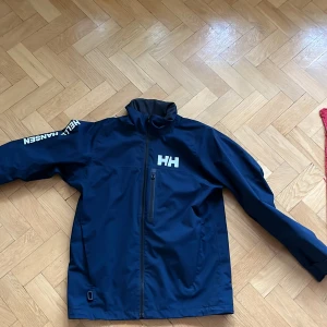 Blå vindjacka från Helly Hansen - Säljer en snygg blå vindjacka från Helly Hansen. Den har en dragkedja framtill och är långärmad med loggan tryckt på ärmen och bröstet. Perfekt för blåsiga dagar och utomhusaktiviteter. Jackan är i bra skick och redo för nya äventyr!