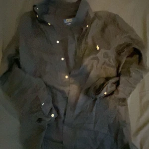 Svart skjorta från Polo Ralph Lauren - Snygg svart skjorta från Polo Ralph Lauren med klassisk krage och knappar hela vägen ner. Den har en broderad logga på bröstet och är perfekt för både vardag och fest. Skjortan är långärmad och gjord i ett bekvämt material. Passar bra till jeans eller chinos för en stilren look. Ny pris för den är 1500 jag säljer för 1200 pris kan diskuteras. Har andvänt den i 2 månader köpt från zalando