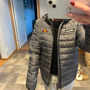 Grå pufferjacka från Ellesse - Säljer en snygg grå pufferjacka från Ellesse. Den är perfekt för kyliga dagar med sin quiltade design och dragkedja framtill. Jackan har en normal passform och är långärmad, vilket gör den idealisk för både höst och vinter. Den har också Ellesse-loggan på bröstet för en sportig look.