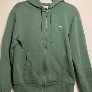 Säljer en snygg grön hoodie från Lacoste i storlek M. Den har en dragkedja framtill och den klassiska krokodilloggan på bröstet. Perfekt för både höst, vinter och vår, med en skön passform och huva för extra mysfaktor. Passar lika bra till vardags som till träning! Tröjan är dock lite mindre än sin storlek. Säljer tröjan eftersom den var för liten. Tröjan har även använts endast ett fåtal gånger men köptes för drygt ett år sedan. Nypris: 1600. Priset är inte hugget i sten!