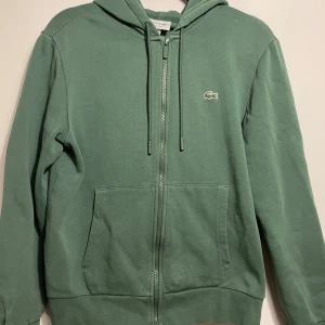 Grön hoodie från Lacoste - Säljer en snygg grön hoodie från Lacoste i storlek M. Den har en dragkedja framtill och den klassiska krokodilloggan på bröstet. Perfekt för både höst, vinter och vår, med en skön passform och huva för extra mysfaktor. Passar lika bra till vardags som till träning! Tröjan är dock lite mindre än sin storlek. Säljer tröjan eftersom den var för liten. Tröjan har även använts endast ett fåtal gånger men köptes för drygt ett år sedan. Nypris: 1600. Priset är inte hugget i sten!