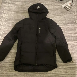Svart pufferjacka från Sail Racing - Säljer en svart pufferjacka från Sail racing i mycket bra skick, haft den i 1,5 år. Jackan har en cool design med loggor på ärmarna och en praktisk huva. Perfekt för kalla vinterdagar med sin varma och vadderade stil. Dragkedja framtill och flera fickor för förvaring. Passar både till vardags och utomhusaktiviteter.
