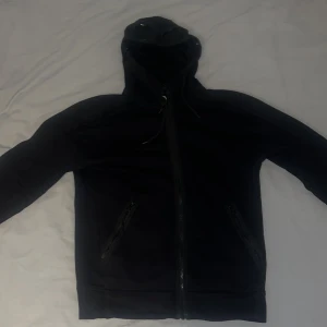 Svart cp company Hoodie med glasögon i huvan - Säljer en cool svart hoodie i storlek M med inbyggda glasögon i huvan. Perfekt för att sticka ut i mängden! Den har dragkedja framtill och långa ärmar. Passar bra för höst och vår. Perfekt för dig som gillar unik stil!