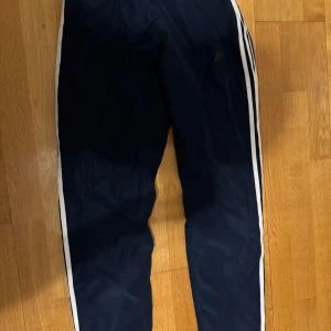 Mörkblå träningsbyxor från Adidas - Säljer ett par mörkblå träningsbyxor från Adidas i bra skick. De har de klassiska vita ränderna längs sidorna och en elastisk midja för extra komfort. Perfekta för träning eller en avslappnad dag. 🏃‍♀️