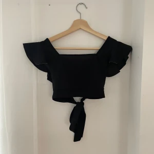 Svart offshoulder topp med knyt - Snygg svart offshoulder topp med volangdetaljer och knyt framtill. Perfekt för sommarens fester eller en utekväll. Toppen är croppad och ger en trendig look.