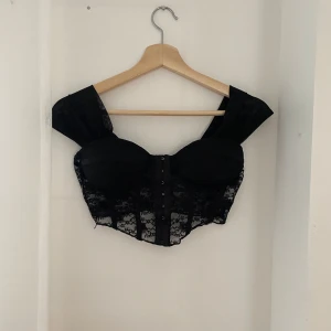 Svart spetskorsett - Snygg svart korsett i spets med vadderade kupor och knäppning fram. Den har en elegant offshoulder-design och är perfekt för festliga tillfällen. Detaljerna i spets ger en lyxig känsla.
